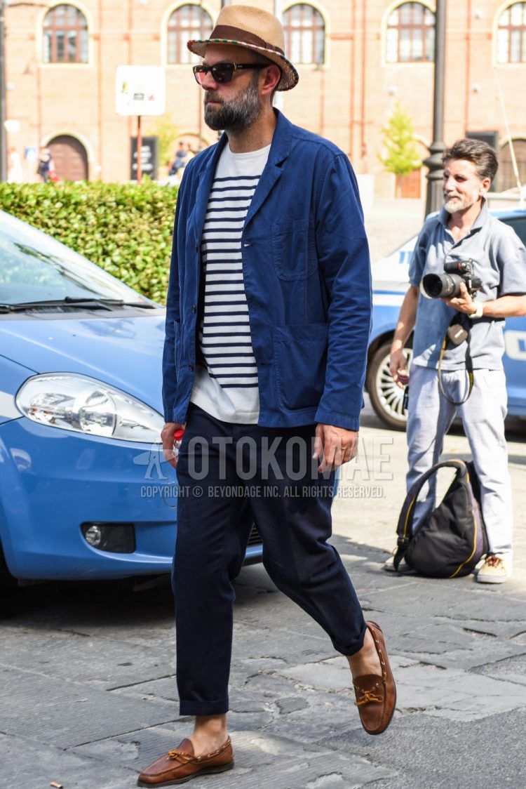 A summer/spring/fall men's coordinate outfit with a solid beige hat, solid blue tailored jacket, white/black striped t-shirt, solid navy cotton pants, and brown moccasin/deck shoe leather shoes.