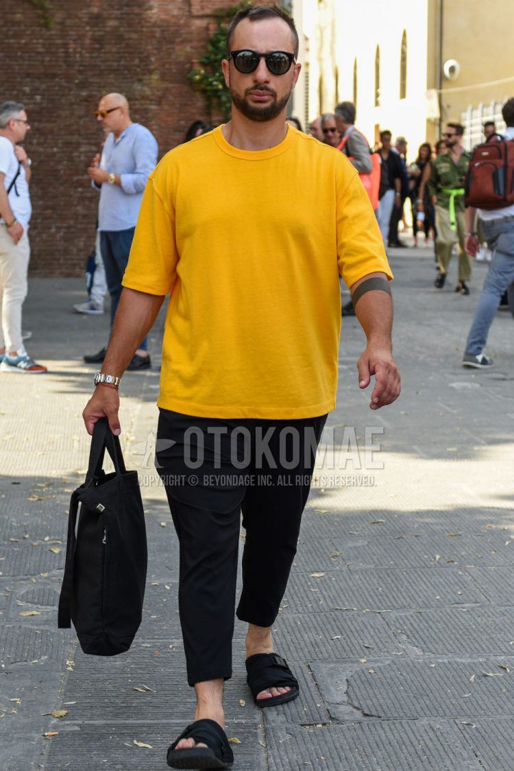 A summer men's coordinate outfit with solid black sunglasses, solid yellow t-shirt, solid black ankle pants, black shower sandals, and solid black tote bag.