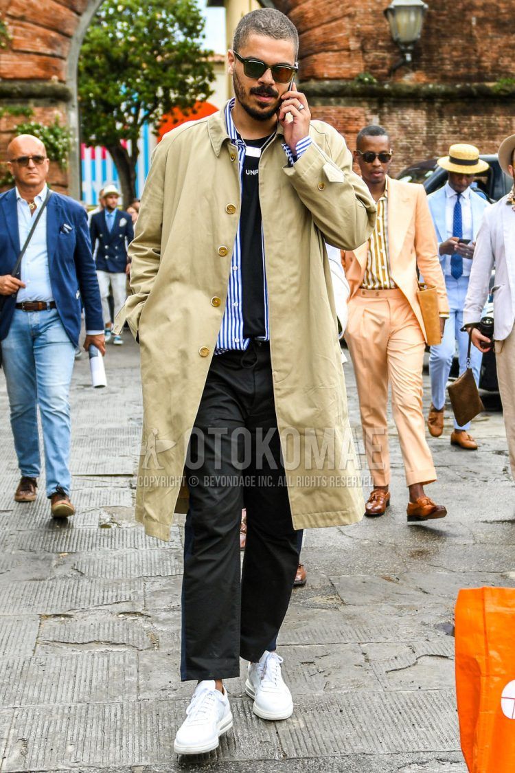 A men's spring/summer/fall outfit for men with plain black sunglasses, plain beige stainless steel collar coat, blue striped shirt, plain navy t-shirt, plain black chinos, and white low-cut sneakers.