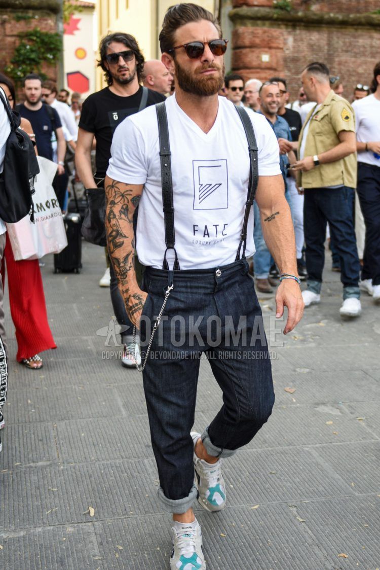 A summer men's coordinate outfit with plain brown sunglasses, white graphic t-shirt, plain black suspenders, plain navy denim/jeans, and white low-cut sneakers.