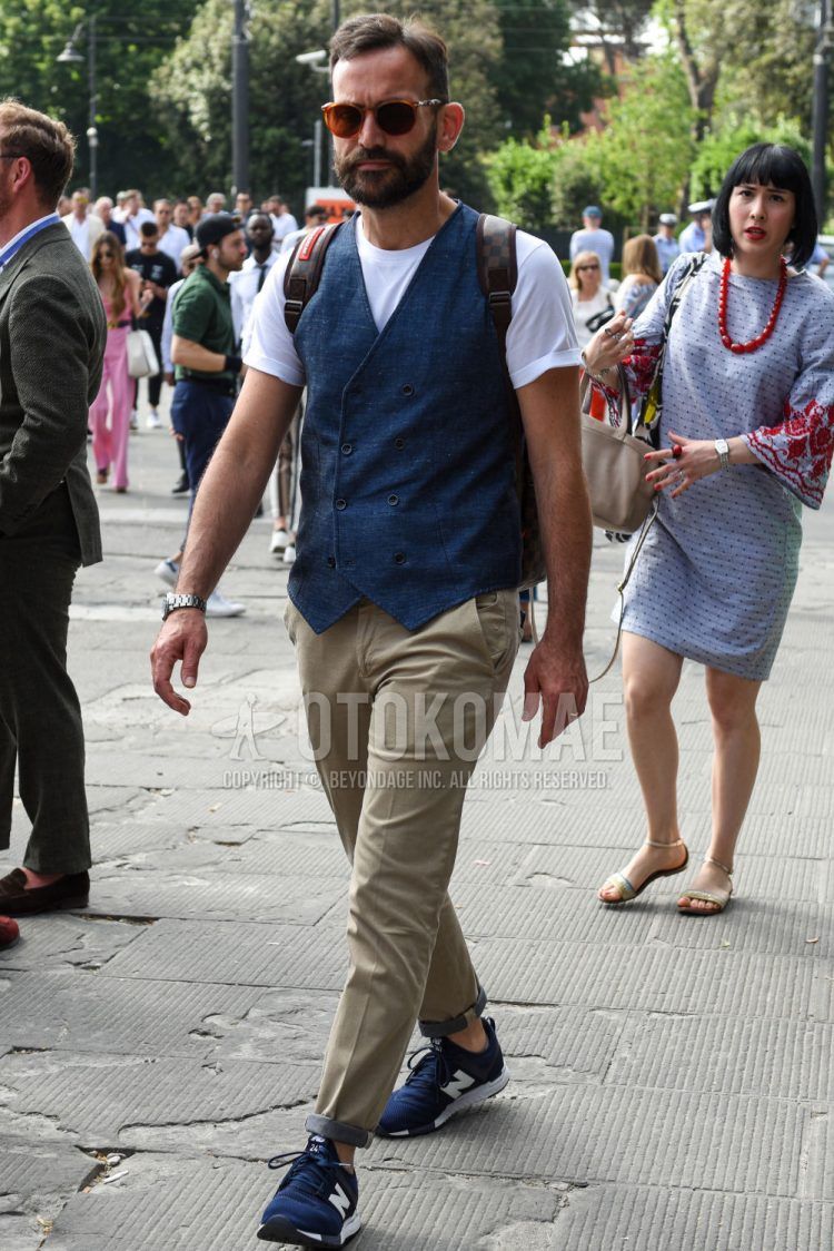 A summer men's coordinate outfit with plain brown sunglasses, plain white t-shirt, plain navy gilet, plain beige cotton pants, and navy low-cut New Balance sneakers.