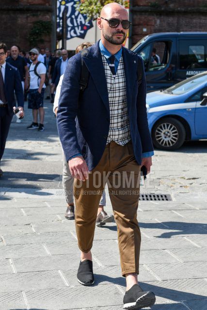 A men's spring/fall outfit with plain black sunglasses, plain navy tailored jacket, plain blue denim/chambray shirt, white checked gilet, plain brown cotton pants, black low-cut sneakers, and navy/white striped knit tie.