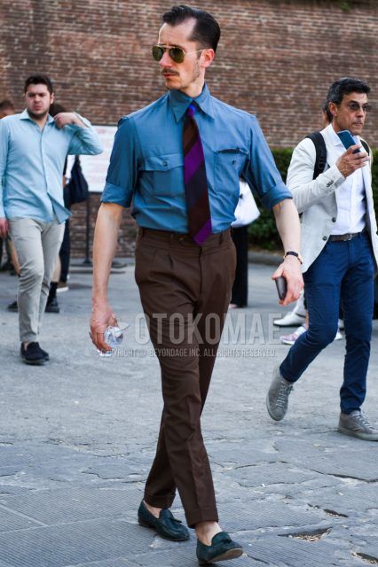 A summer/spring men's outfit with teardrop black and gold solid sunglasses, solid light blue shirt, solid brown beltless pants, black tassel loafer leather shoes, and navy regimental tie.