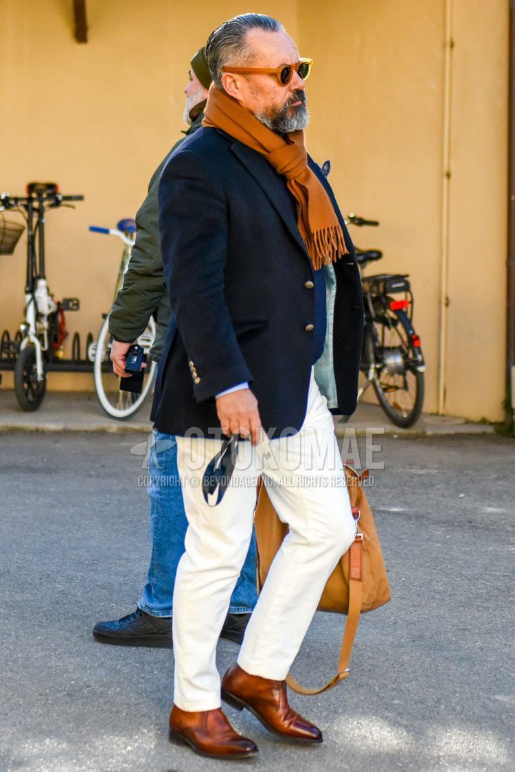 A men's fall/winter outfit with solid color sunglasses, solid color brown scarf/stall, solid color navy tailored jacket, solid color white slacks, and brown chukka boots.