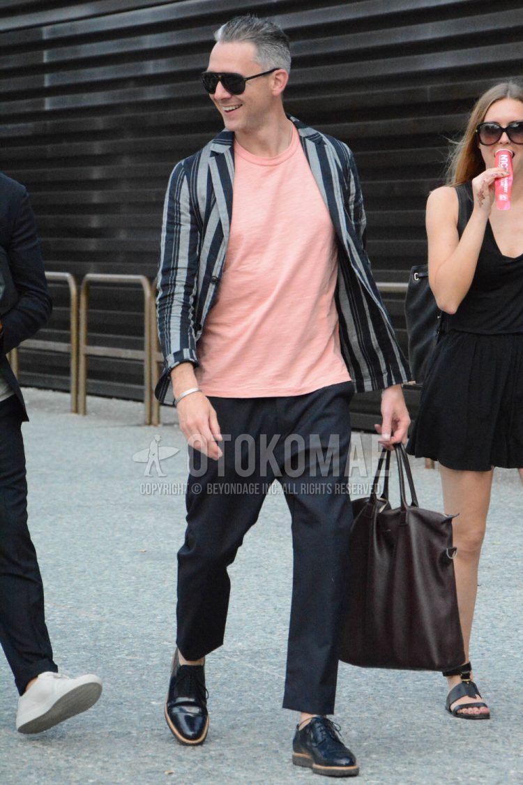 A men's spring/summer/fall outfit with plain black sunglasses, black/gray striped tailored jacket, plain pink t-shirt, plain black slacks, black straight tip leather shoes, and plain brown tote bag.