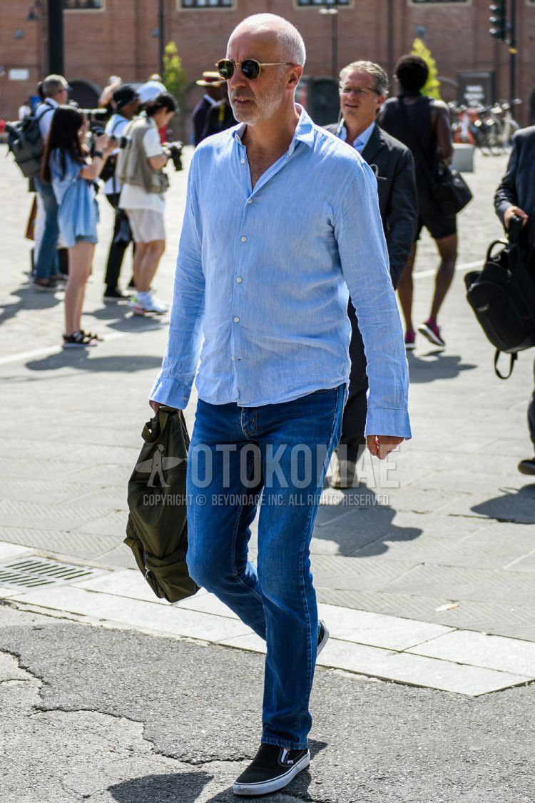 Spring/summer men's coordinate outfit with solid beige sunglasses, solid linen light blue shirt, solid blue denim/jeans, and black slip-on sneakers.