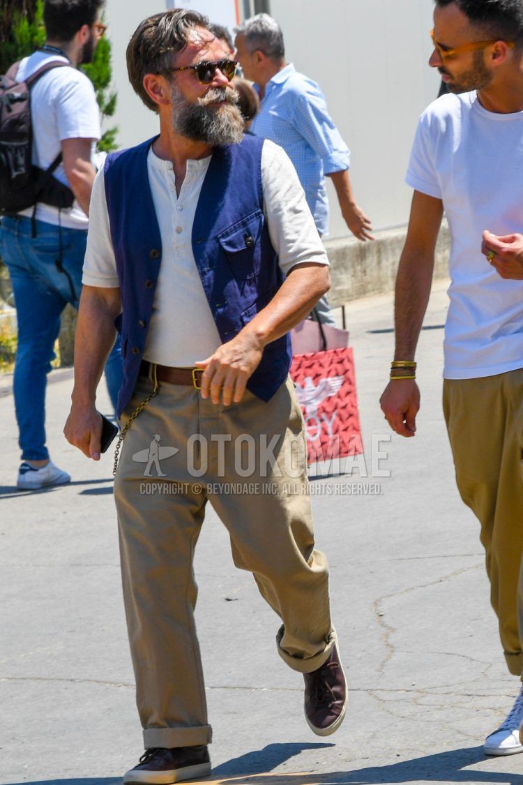 A spring/summer men's coordinate outfit with plain sunglasses, a plain T-shirt with a henley neck, a plain navy gilet, a plain leather belt, plain beige chinos, and brown low-cut sneakers.