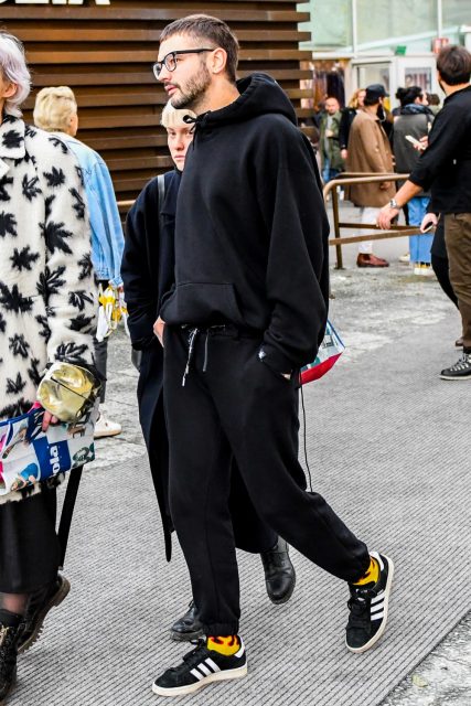 Winter men's oversized coordinate outfit with solid color glasses, solid color black hoodie, solid color black sweatpants, solid color yellow socks, and black low-cut Adidas sneakers.