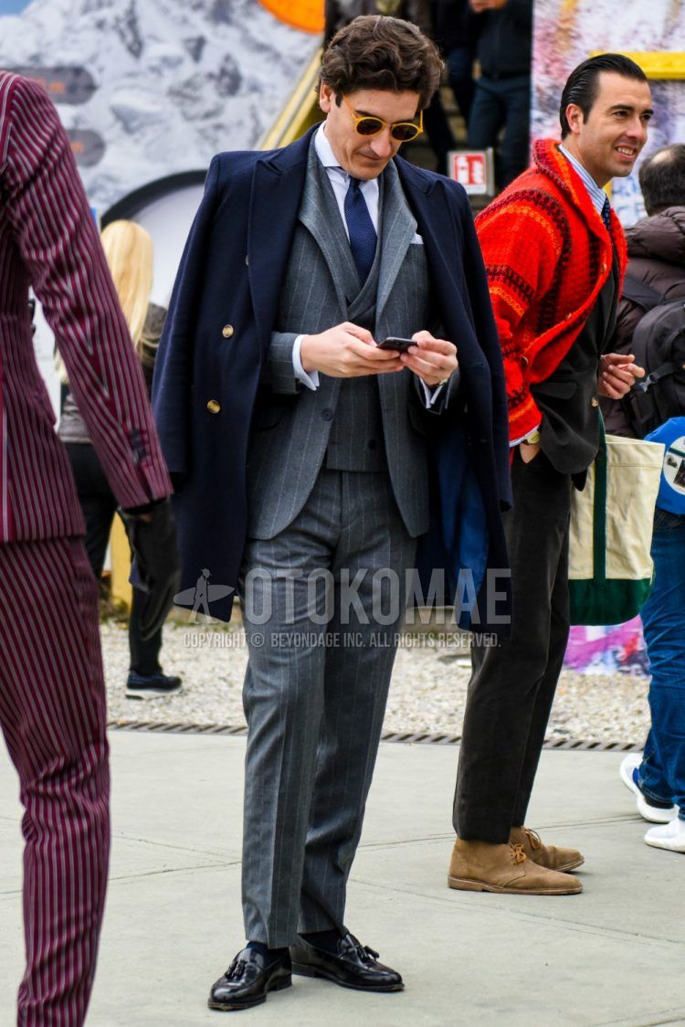Fall and winter men's outfit with plain yellow sunglasses, plain navy Ulster coat, plain white shirt, plain navy socks, black tassel loafer leather shoes, gray striped three-piece suit, and plain navy tie.