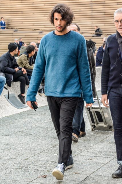 A men's oversized fall/spring outfit with a solid light blue sweater, solid gray slacks, and black high-cut sneakers.