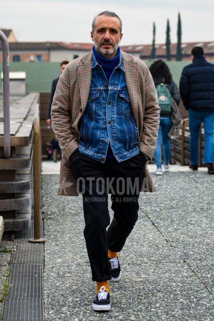 A men's fall/winter outfit with a solid blue denim jacket, beige checked stainless steel coat, solid blue turtleneck knit, solid black wool pants, solid yellow socks, and black low-cut sneakers from Vans Authentic.
