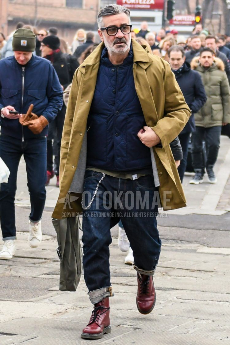 Winter men's outfit with solid color glasses, solid color beige stainless coat, solid color navy quilted jacket, solid color navy denim/jeans, Dr. Martens red other boots, and solid color gray tote bag.