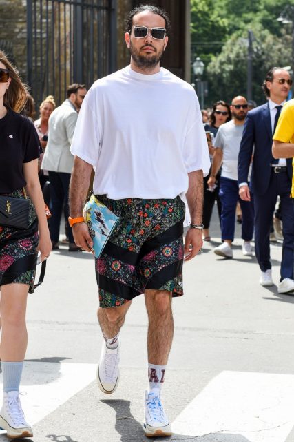 A summer men's oversized coordinate outfit with plain white sunglasses, plain white t-shirt, multi-colored and other shorts, plain white socks, and white low-cut sneakers.