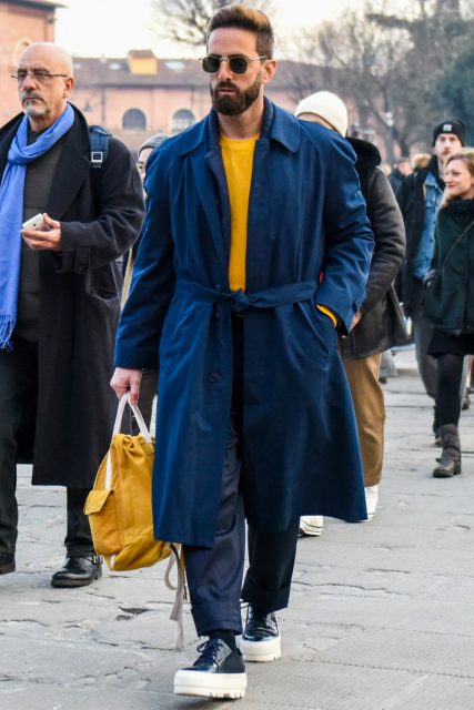 A winter men's oversized coordinate outfit with solid color sunglasses, solid color navy belted coat, solid color yellow sweatshirt, solid color navy cotton pants, solid color navy socks, navy low-cut sneakers, and solid color yellow Boston bag.