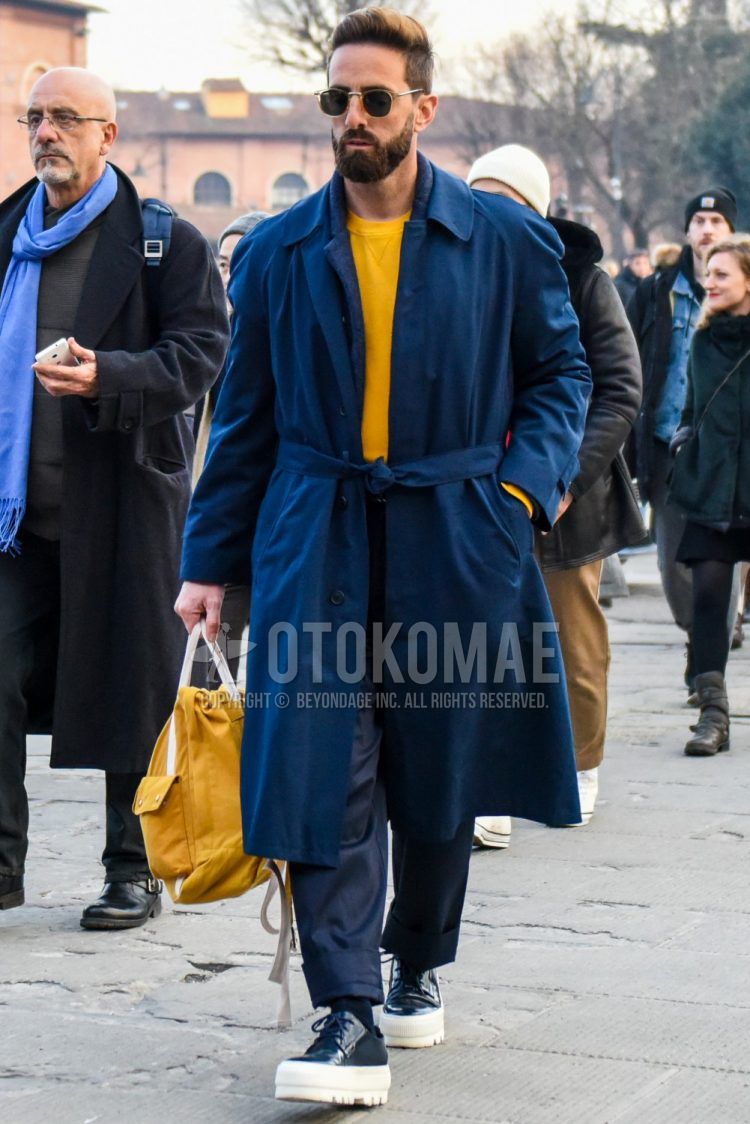 A winter men's outfit outfit with solid color sunglasses, solid color navy belted coat, solid color yellow sweatshirt, solid color navy cotton pants, solid color navy socks, navy low-cut sneakers, and solid color yellow Boston bag.