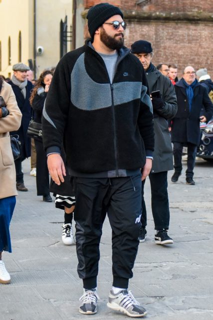 Winter men's oversized coordinate outfit with solid black knit cap, solid sunglasses, solid black fleece jacket, solid gray sweater, solid black jogger pants/ribbed pants by Nike, solid white socks, and white low-cut sneakers by Adidas.