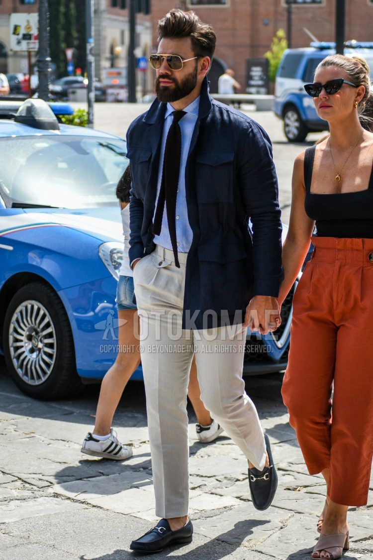 A spring and fall men's outfit with plain gold sunglasses, plain navy M-65, plain light blue shirt, plain white beltless pants, navy bit loafer leather shoes, and plain navy knit tie.