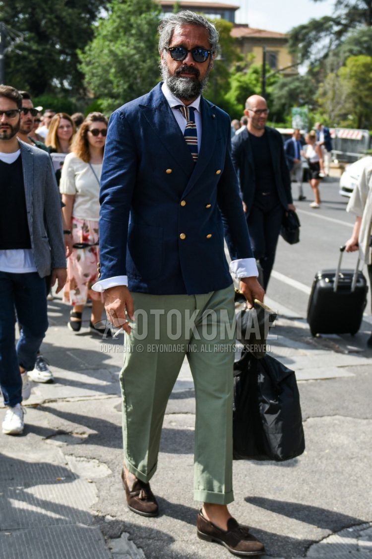 A spring/fall men's outfit with beige/black tortoiseshell sunglasses, plain navy tailored jacket, plain white shirt, plain green chinos, suede brown tassel loafer leather shoes, plain black briefcase/handbag, and navy striped knit tie.