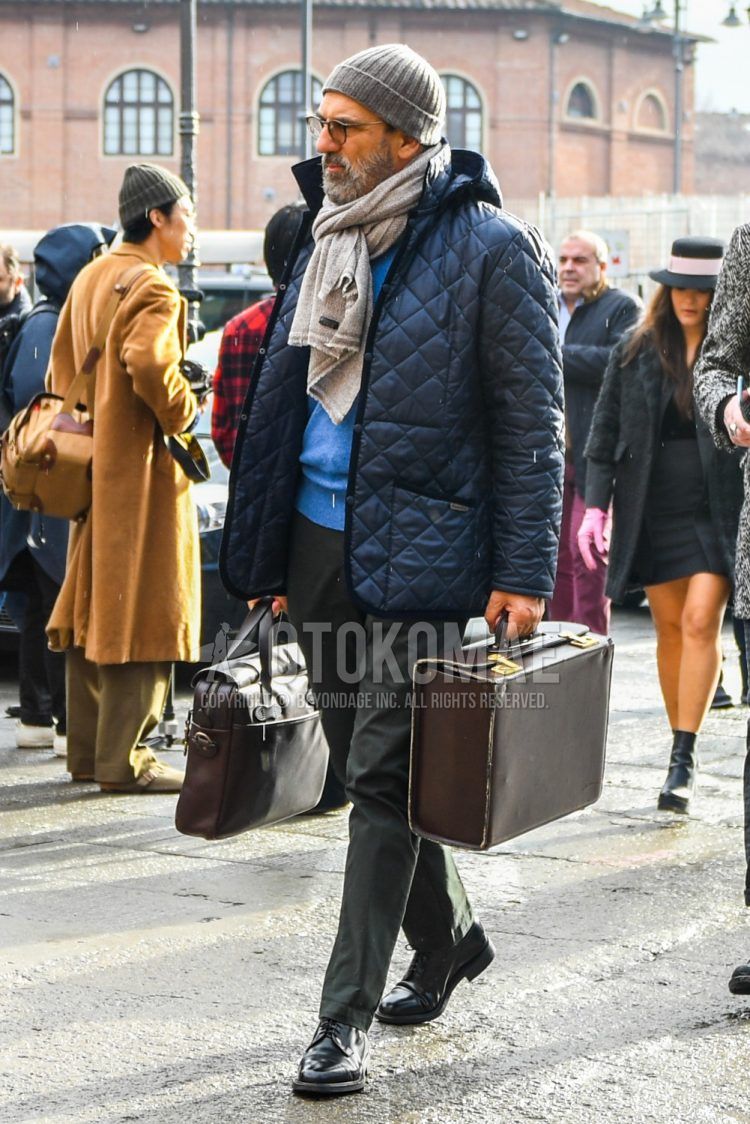 Men's fall/winter outfit with plain gray knit cap, solid color glasses, solid beige scarf/stall, solid navy quilted jacket, solid blue sweater, solid gray cotton pants, black straight tip leather shoes, solid brown briefcase/handbag outfits.
