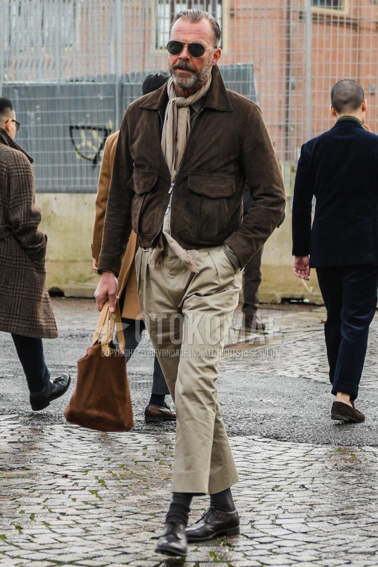 A winter men's outfit with solid color sunglasses, solid color brown scarf/stall, solid color brown leather jacket (other than rider's), solid color beige pleated pants, solid color dark gray socks, and brown plain toe leather shoes.
