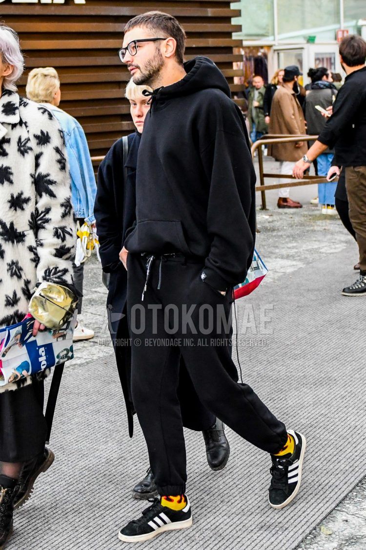 Men's coordinate and outfit with solid color glasses, solid color black hoodie, solid color black sweatpants, solid color yellow socks, and black low-cut Adidas sneakers.