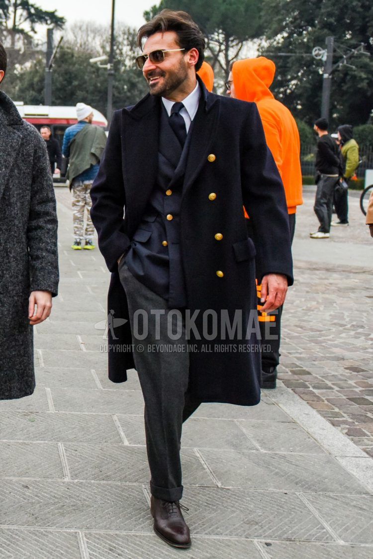 A winter men's outfit with plain sunglasses, plain navy chester coat, plain navy tailored jacket, dark gray plain wool slacks, and brown brogue shoes leather shoes.