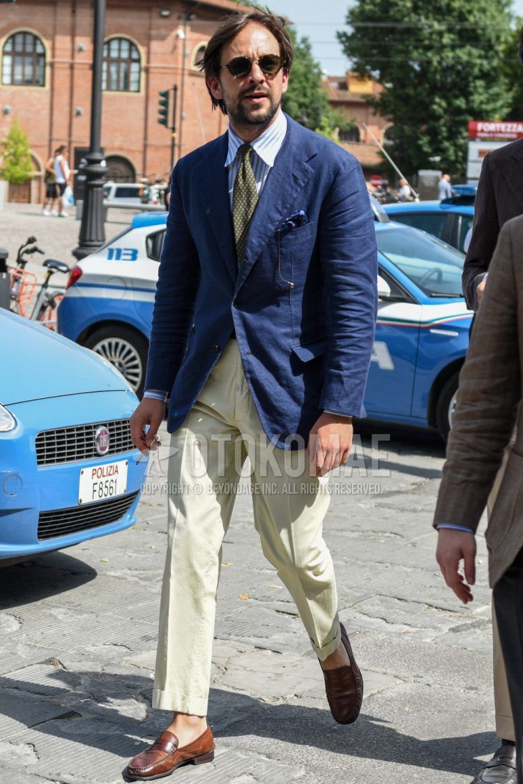 Spring, summer and fall men's coordinate outfit with plain black sunglasses, plain navy tailored jacket, white and light blue striped shirt, plain beige cotton pants, brown coin loafer leather shoes and olive green dot tie.