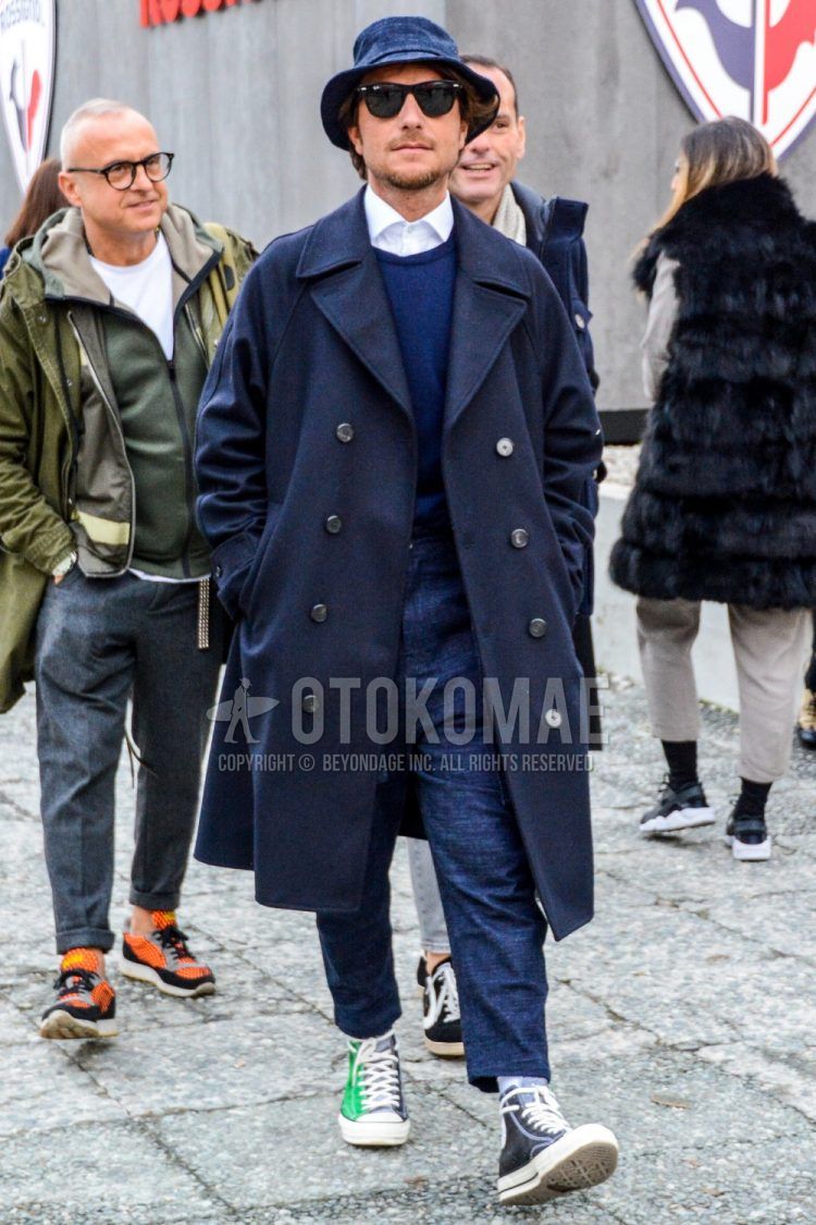 A men's fall/winter outfit with a solid hat, solid sunglasses, solid navy Ulster coat, solid navy sweater, solid white shirt, solid gray slacks, solid white socks, and Converse All Star black/green high-cut sneakers.