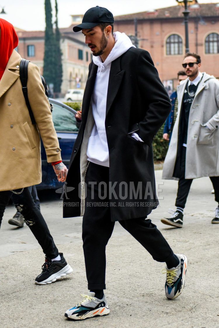 A men's fall/winter outfit with a solid black baseball cap, solid black chester coat, solid white hoodie, solid black slacks, solid black socks, and Adidas Yeezy Boost 700 gray low-cut sneakers.