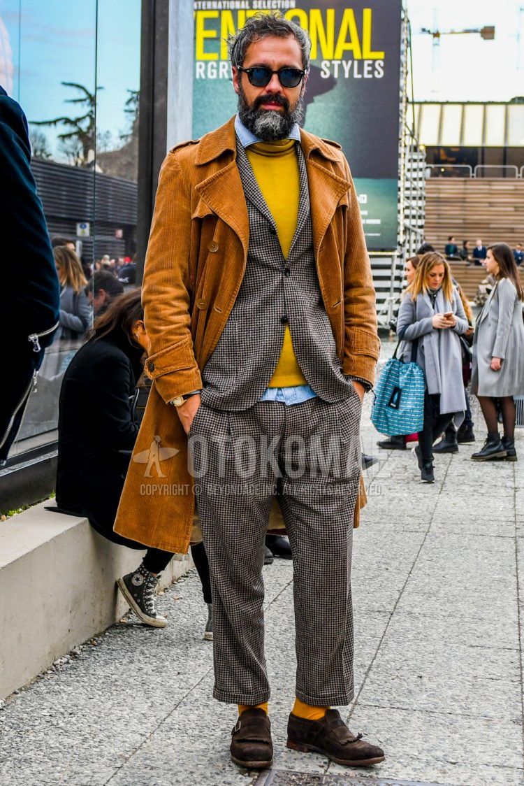 Fall/winter men's outfit with solid color sunglasses, solid color beige trench coat, solid color yellow turtleneck knit, solid color blue denim/chambray shirt, solid color yellow socks, brown monk shoes leather shoes, brown suede shoes leather shoes.