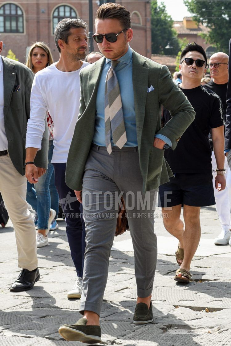 A spring/fall men's outfit with solid black sunglasses, solid olive green tailored jacket, solid blue denim/chambray shirt, solid gray slacks, olive green slip-on sneakers, and a multi-colored regimental tie.