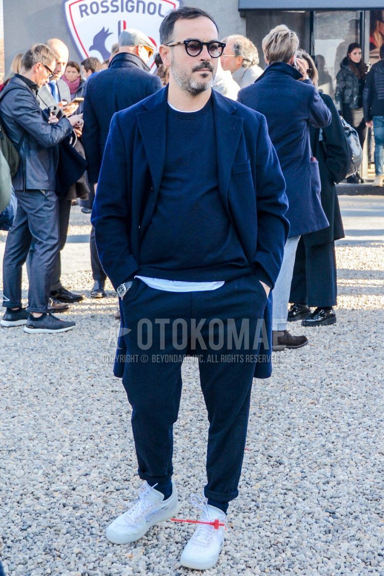 A men's fall/winter outfit with plain sunglasses, plain navy chester coat, plain navy sweater, plain white t-shirt, plain navy cotton pants, plain navy socks, and white low-cut sneakers.