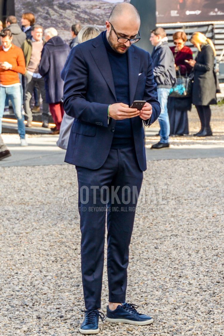 A men's fall outfit with plain glasses, a plain navy turtleneck knit, black low-cut sneakers, and a plain gray suit.