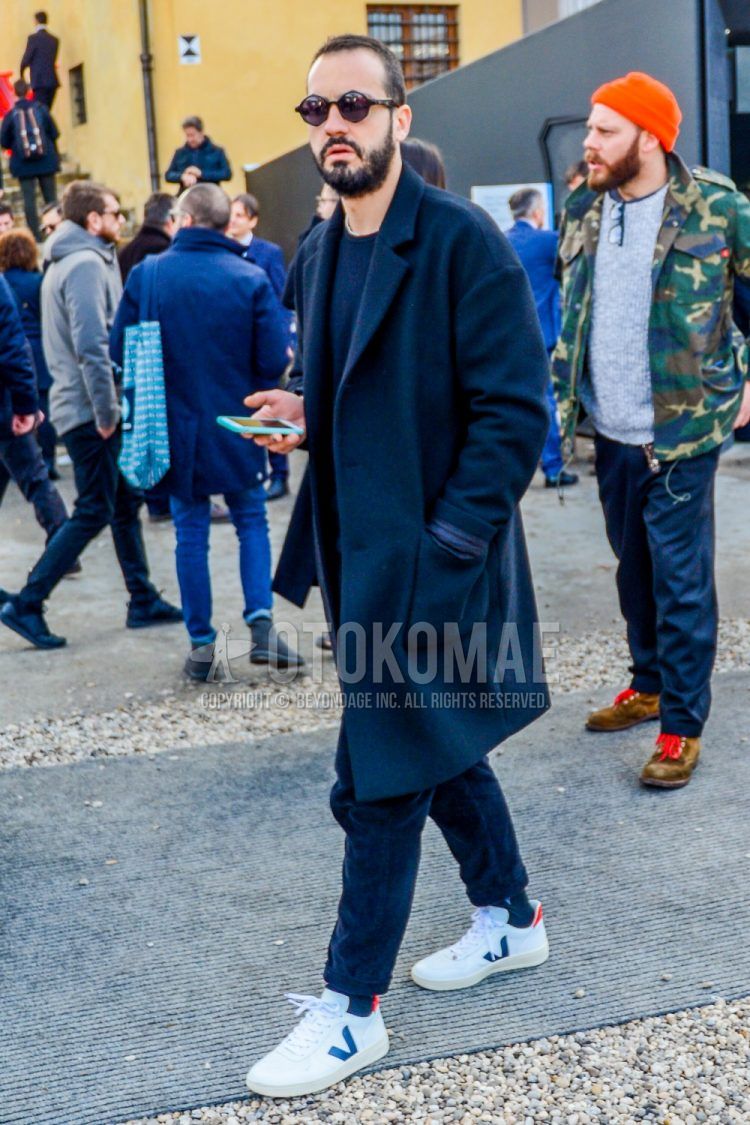 A men's fall/winter outfit with plain sunglasses, plain navy chester coat, plain navy sweater, plain navy chinos, plain navy socks, and white low-cut sneakers.
