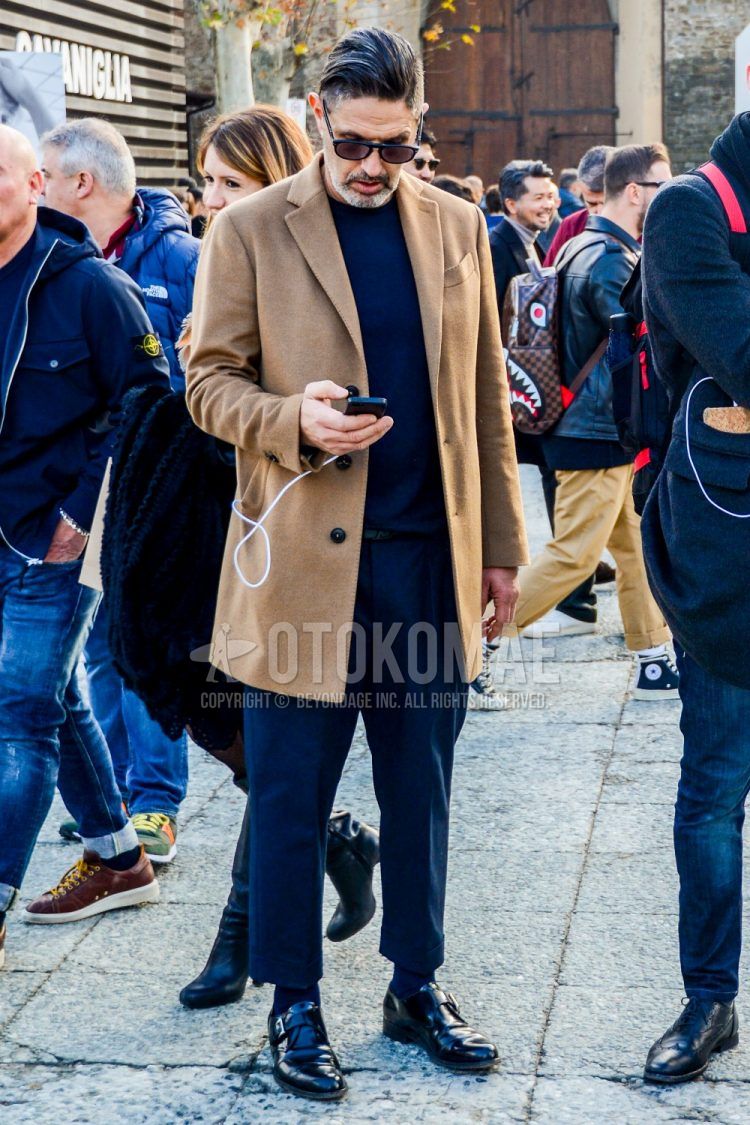 A men's fall/winter outfit with solid color sunglasses, solid color beige chester coat, solid color navy sweater, solid color gray slacks, solid color navy socks, and black monk leather shoes.