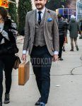 Men's outfit/clothing with brown checked tailored jacket, brown checked gilet, solid navy bottoms, solid blue socks, black coin loafer leather shoes, and solid beige briefcase/handbag.