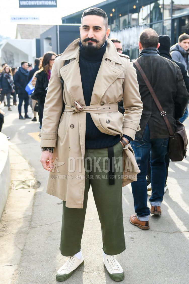 Winter men's coordinate outfit with plain beige trench coat, plain navy turtleneck knit, plain olive green wide pants, plain olive green chinos, and white/olive green low-cut sneakers.