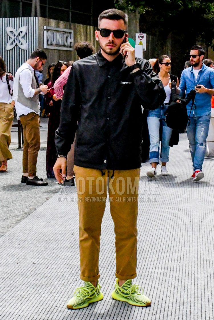 A summer/fall/spring men's coordinate outfit with solid color sunglasses, solid color black coach jacket, solid color beige chinos, and easy boost 350 semi-frozen yellow yellow low-cut sneakers.