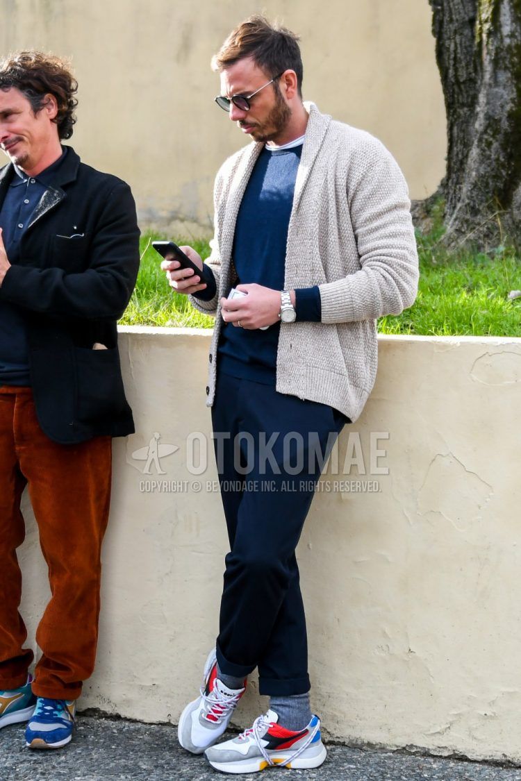A winter men's coordinate outfit with solid black sunglasses, solid beige cardigan, solid navy sweater, solid navy slacks, solid gray socks, and Diadora multi-colored low-cut sneakers.