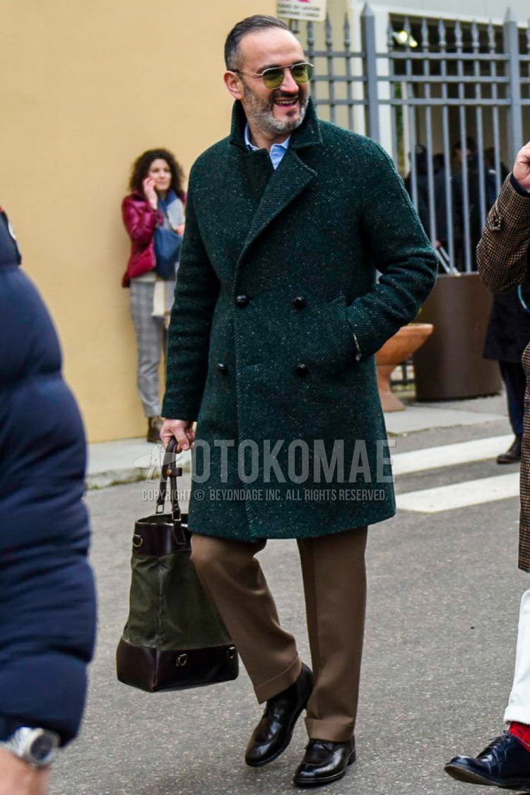Winter men's outfit with solid color sunglasses, green herringbone chester coat, solid color brown slacks, brown U-tip leather shoes, and solid color brown/olive green tote bag.