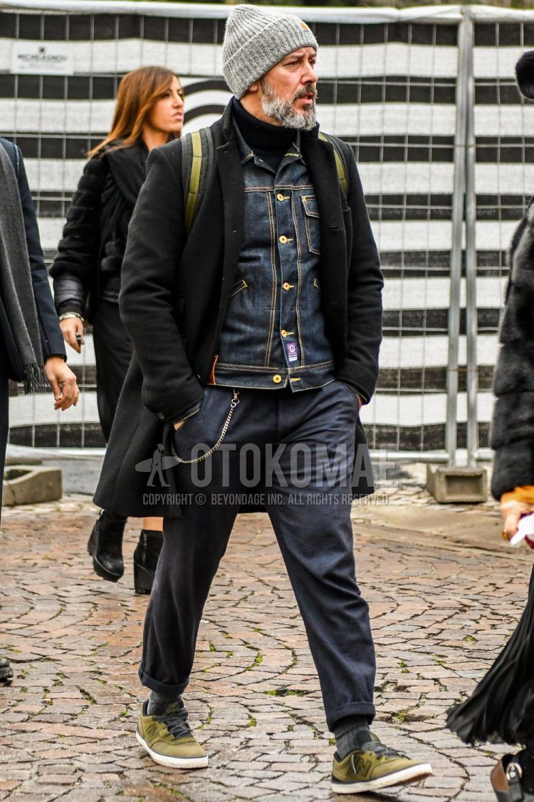 A men's fall/winter outfit with a solid gray knit cap, solid black chester coat, solid navy denim jacket, solid black turtleneck knit, solid gray slacks, solid gray socks, and New Balance olive green low-cut sneakers.
