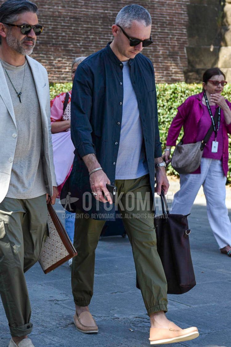 A spring/summer/fall men's outfit with solid black sunglasses, solid black shirt, solid gray t-shirt, solid olive green jogger pants/ribbed pants, beige moccasins/deck shoes leather shoes, and solid brown tote bag.
