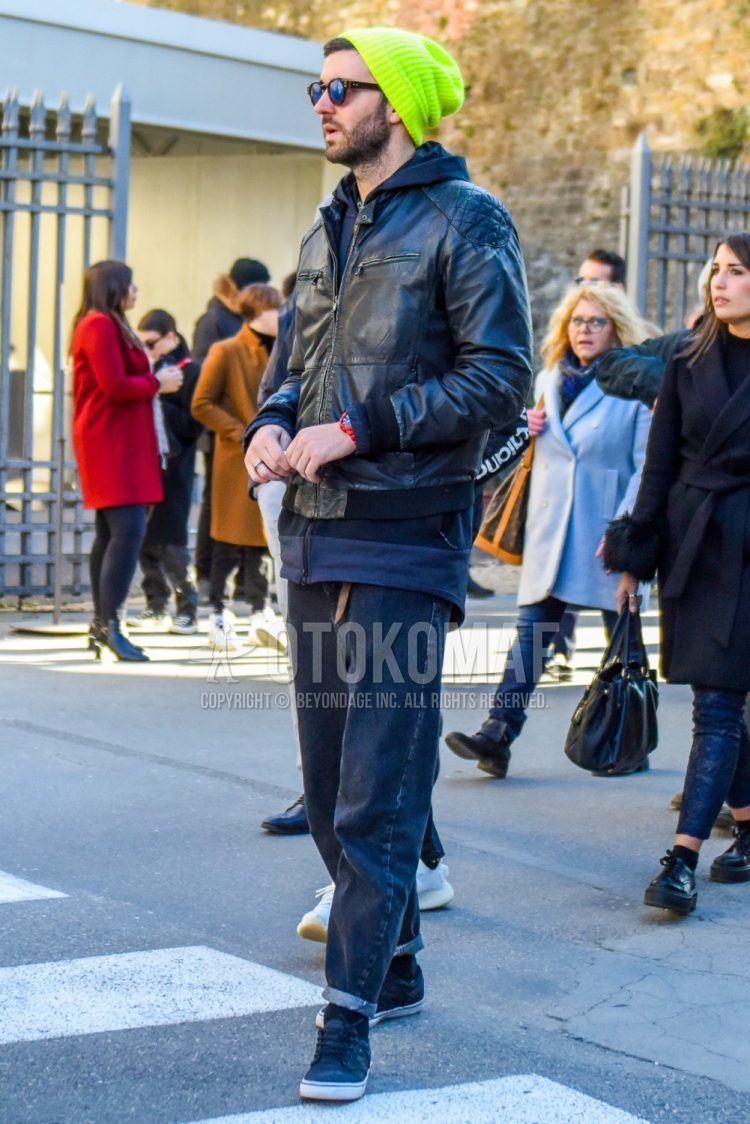 Fall/winter men's outfit with plain yellow knit cap, plain black sunglasses, plain black rider's jacket, dark gray/black plain hoodie, plain leather belt, dark gray plain denim/jeans, plain black socks, and dark gray low-cut sneakers.