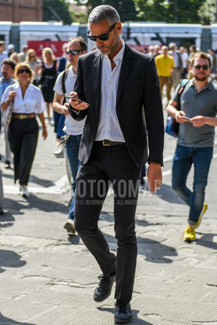 A spring and fall men's coordinate outfit with plain black sunglasses, a plain white shirt, a plain black leather belt, black low-cut sneakers, and a plain black suit.