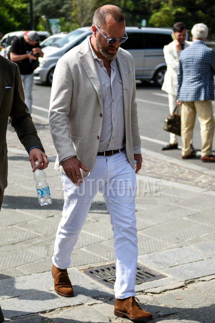 A spring and fall men's outfit with clear solid color sunglasses, solid color beige tailored jacket, solid color gray/beige shirt, solid color black leather belt, solid color white cotton pants, and suede brown chukka boots.