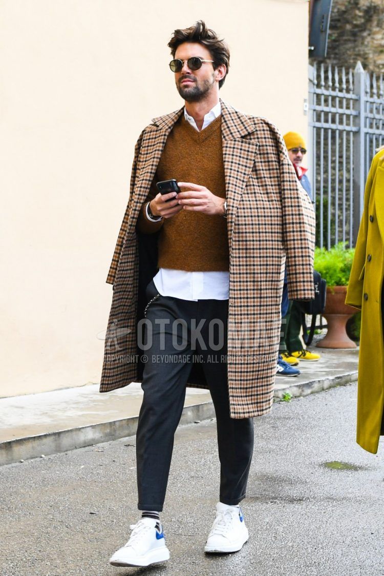 A men's fall/winter outfit with plain sunglasses, brown checked stainless steel coat, plain brown sweater, plain white shirt, plain gray slacks, plain white socks, and white low-cut sneakers by Alexander McQueen.