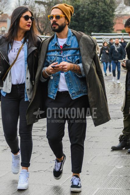 A winter men's outfit wearing a solid brown knit cap, solid sunglasses, solid blue denim jacket, solid brown hooded coat, solid white t-shirt, solid black ankle pants, and black low-cut sneakers by Vans.