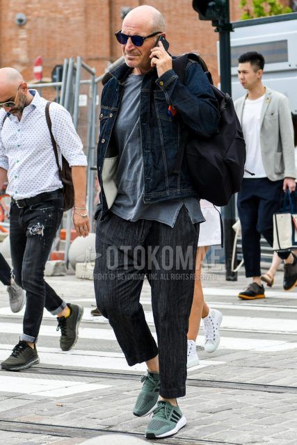 A spring/summer/fall men's coordinate outfit with solid black sunglasses, solid navy denim jacket, solid dark gray t-shirt, solid navy denim/jeans, Adidas green low-cut sneakers, and solid black backpack.