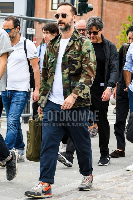 Men's coordinate and outfit with plain sunglasses, multi-colored camouflage M-65, plain white t-shirt, plain navy chinos, Nike beige low-cut sneakers, and plain olive green briefcase/handbag.