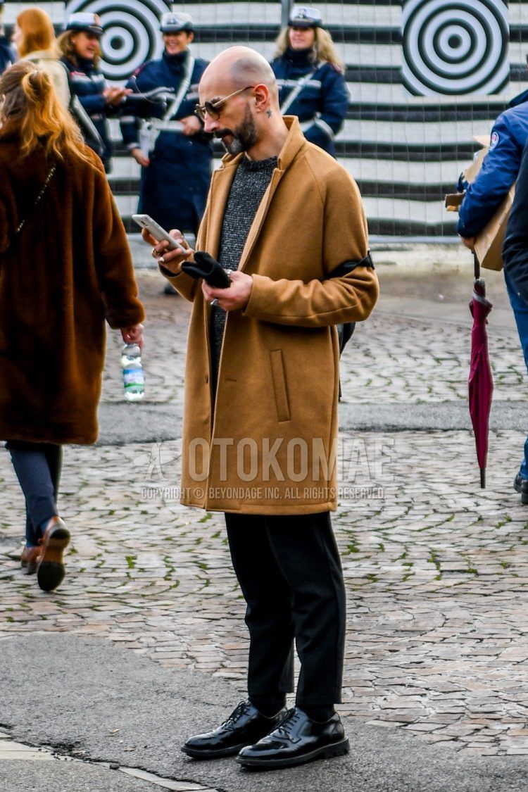 A men's fall/winter outfit with solid color sunglasses, solid color beige chester coat, solid color black sweater, solid color black slacks, solid color black socks, and black plain toe leather shoes.